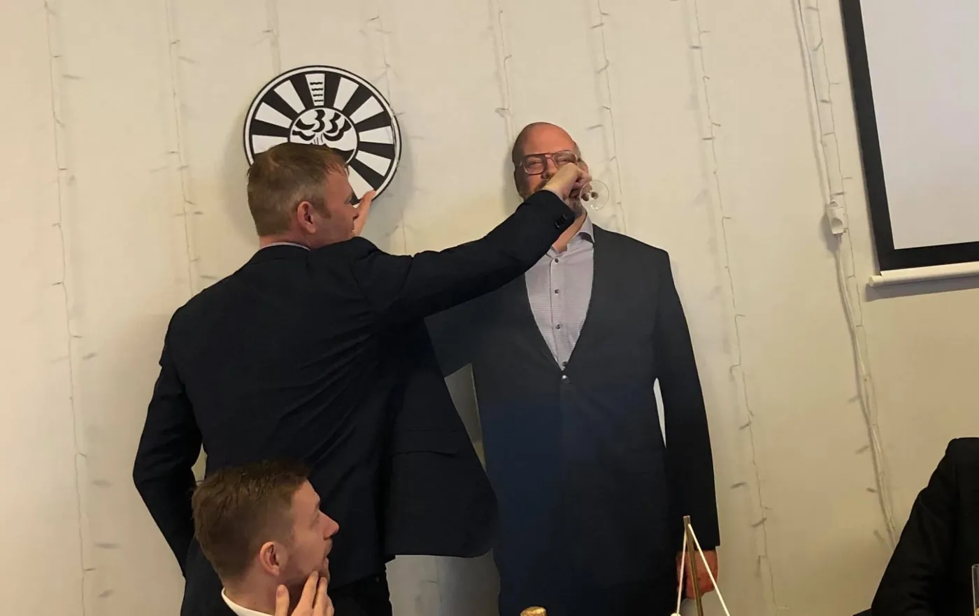 Félagi gefur öðrum að drekka úr glasi fyrir framan Round Table merkið